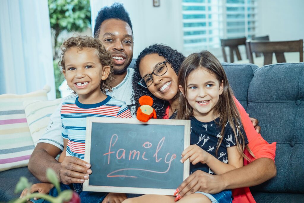 Smiling family of four indoors holding a blackboard with 'family' written on it.